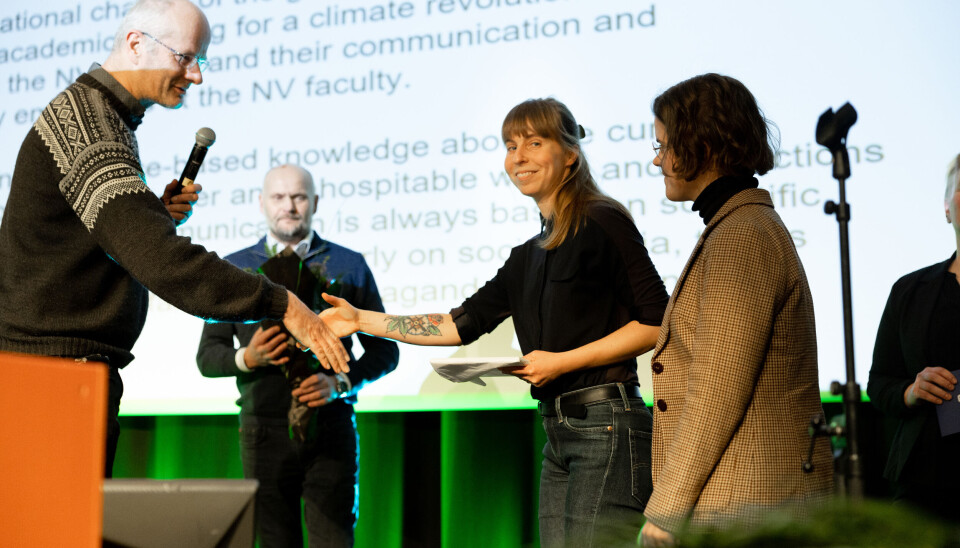 Postdoktor Kristiina Visakorpi (midten) og stipendiat Angeline Bruls mottar prisen Communicator of the Year på vegne av Scientist Rebellion Norway, fra dekan ved Fakultet for naturvitenskap Øyvind Gregersen. Øyvind Gregersen, i strikkegenser, holder ut hånda til Kristiina Visakorpi, en kvinne med svart genser og brunt hår som smiler mot kamera. De står på en scene med en skjerm full av skrift i bakgrunnen.